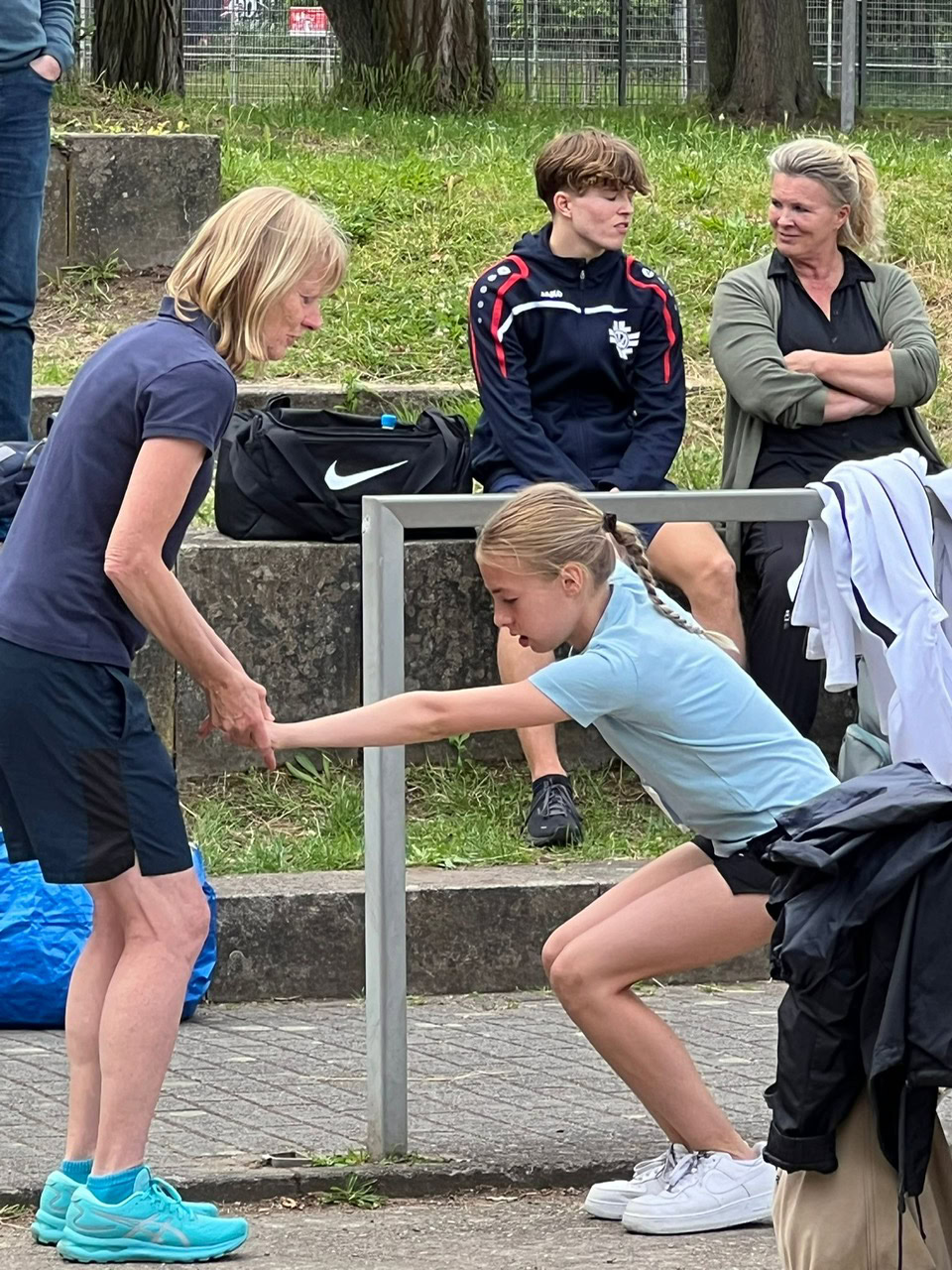 Trainerin hilft einer jungen Sportlerin beim Dehnen vor einem Training oder Wettkampf bei TSG Sonnenberg. Im Hintergrund sitzen weitere Betreuer und Sportler auf der Sitzbank und bereiten sich vor. Die Szene zeigt Gemeinschaft und Engagement im Vereinssport.