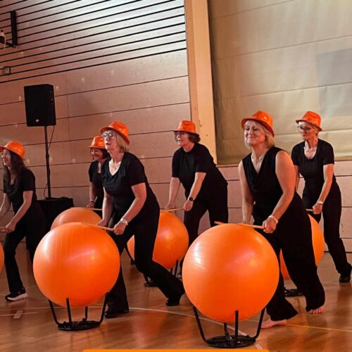 Aktive Frauen beim Drummen mit orangefarbenen Gymbällen und Hüten, auftreten bei einem Event im TSG Sonnenberg, sportliche und unterhaltsame Darbietung in der Sporthalle.