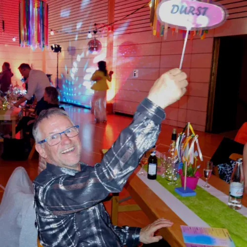 TSG Sonnenberg Feier, Mann hält "Durst" Schild, bunte Dekoration, festliche Veranstaltung, gesellige Atmosphäre, Gemeinschaft, Indoor-Event, festliches Ambiente, Spaß und Zusammenkunft.